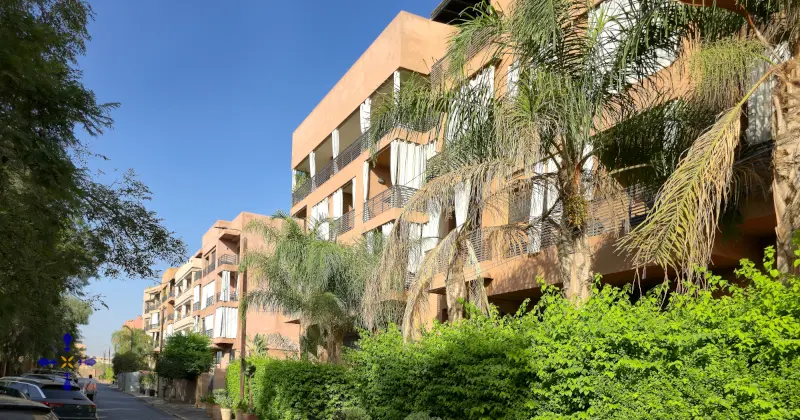 Résidence sécurisée prestige Hivernage Marrakech 2026 : immeubles modernes terracotta, balcons rideaux blancs, palmiers et végétation luxuriante sous ciel bleu, ambiance calme retraité français non-résident